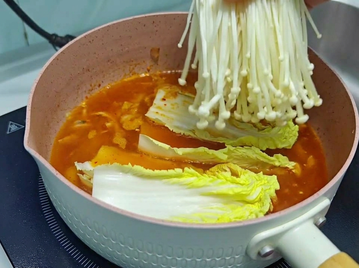 Un primo piano di una mano che cala un mazzetto di funghi enoki frescos e puliti in una pentola rosa contenente una ricca base di zuppa di kimchi rosso-arancio e cavolo cinese.