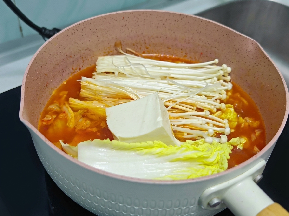 Un grande pezzo di tofu bianco fresco adagiato sopra funghi enoki e cavolo allinterno di una pentola di zuppa di kimchi arancione.