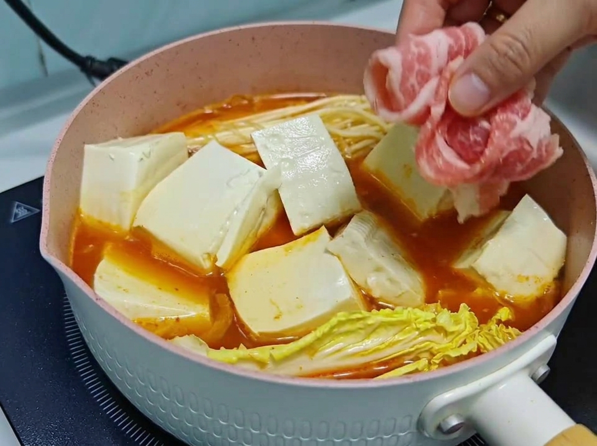 Una mano che adagia con cura sottili fettine di manzo crudo arrotolate sopra cubetti di tofu e verdure in una pentola di zuppa di kimchi che sobbolle.