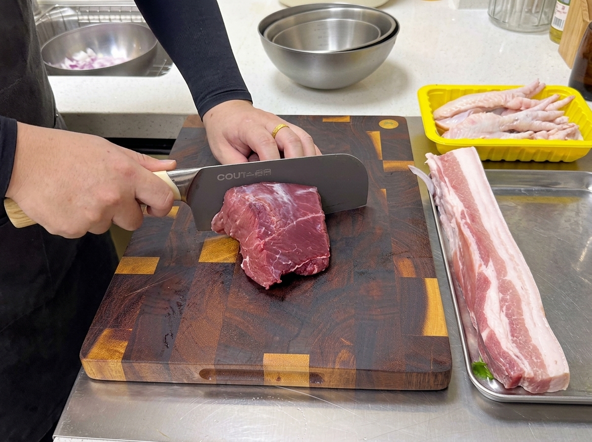 Un cuoco usa un grande coltello professionale per tagliare un pezzo di sottopesce di manzo crudo su un tagliere di legno.