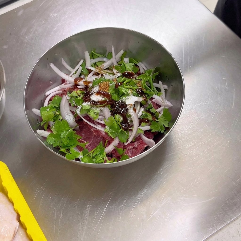 Una ciotola di metallo contenente fette di manzo crudo, cipolle e coriandolo sormontata da spezie secche tra cui cumino in polvere, semi di cumino, sale e zucchero.