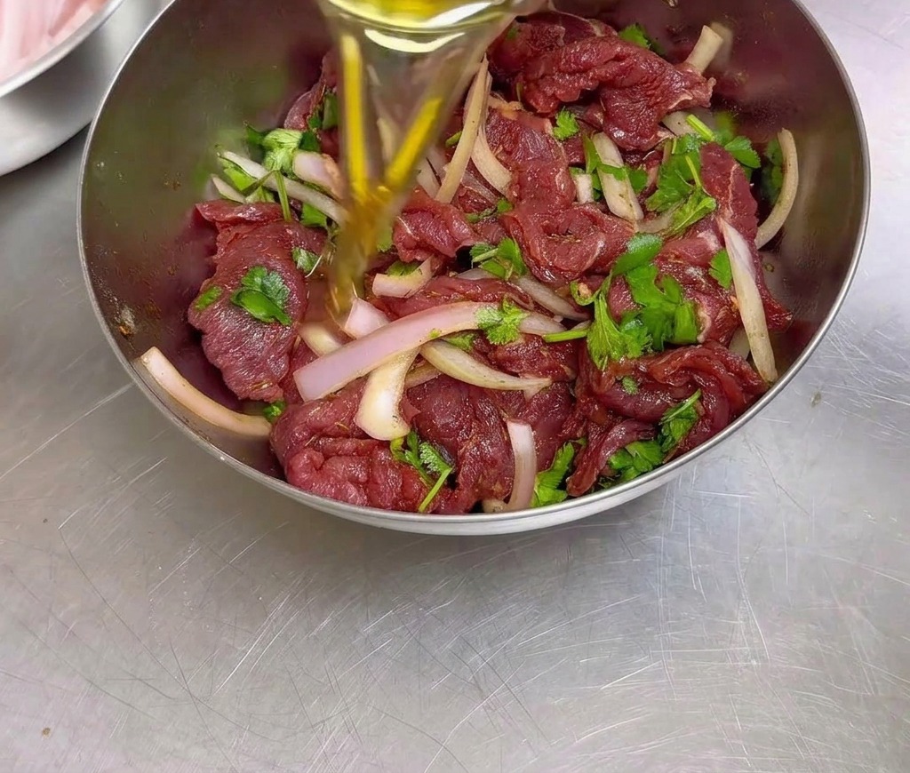 Una persona versa olio di semi in una ciotola di acciaio inossidabile riempita con manzo condito in marinatura, cipolle affettate e coriandolo.