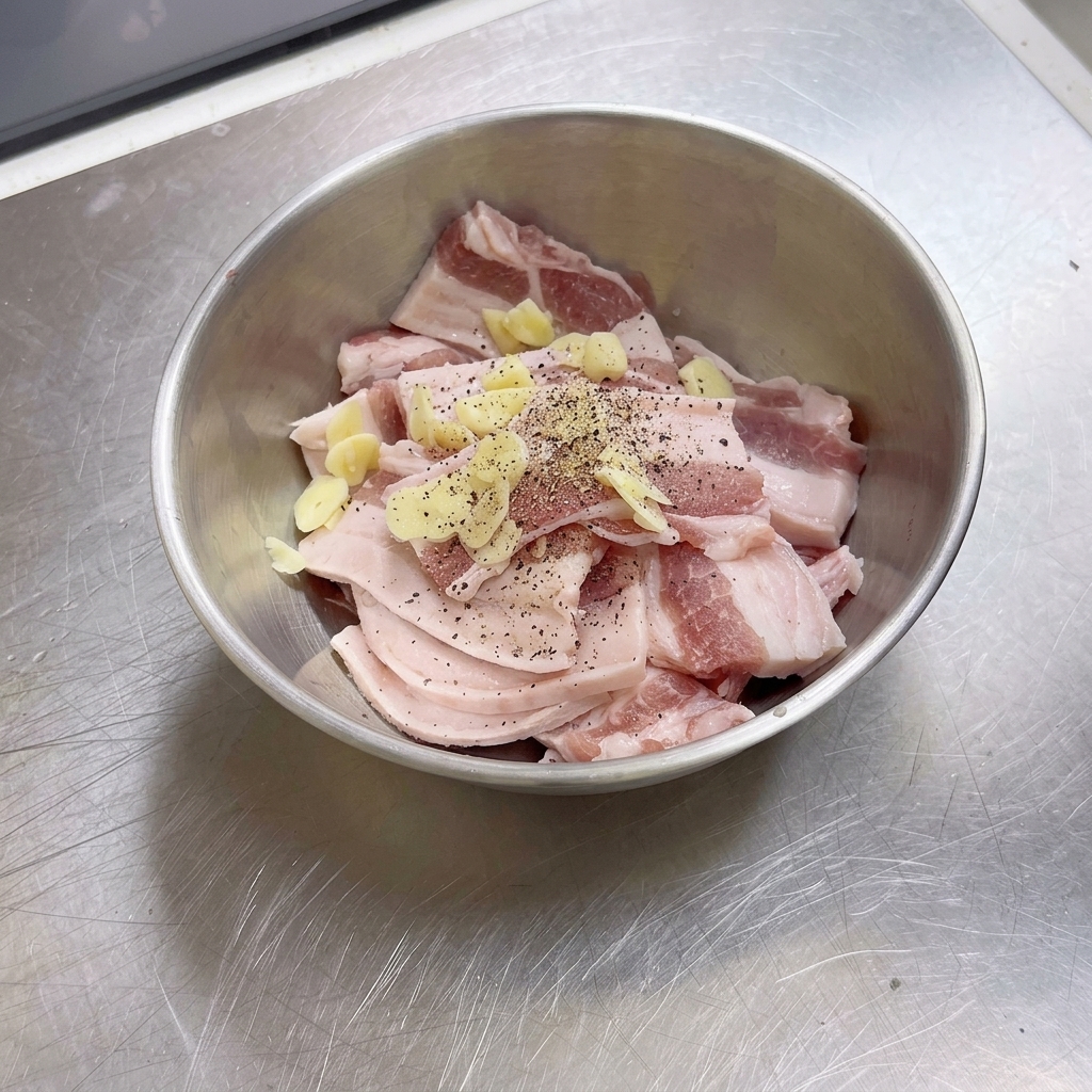 Una ciotola di acciaio inossidabile riempita con fette di pancetta di maiale cruda, pezzi daglio e pepe nero appena macinato.