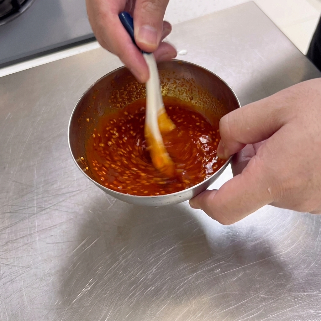 Una densa salsa barbecue di colore rosso scuro ricca di semi di sesamo viene mescolata con un cucchiaio in una piccola ciotola di acciaio inossidabile.