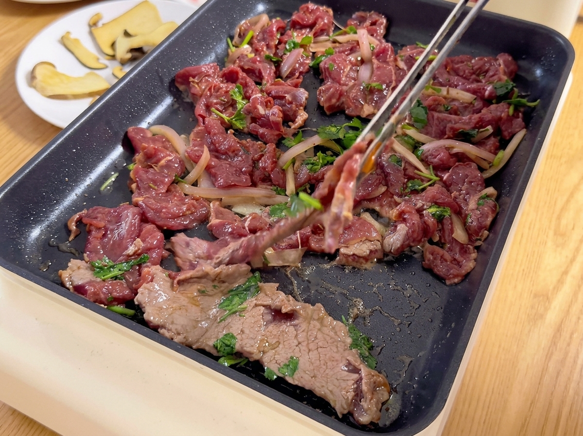 Sottili fette di manzo marinato mescolate con cipolle e coriandolo vengono adagiate su una piastra elettrica calda con laiuto di pinze.