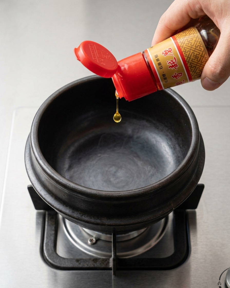 Olio di sesamo profumato che viene versato da una bottiglia di vetro con tappo rosso in una pentola di pietra nera vuota sul fornello a gas.