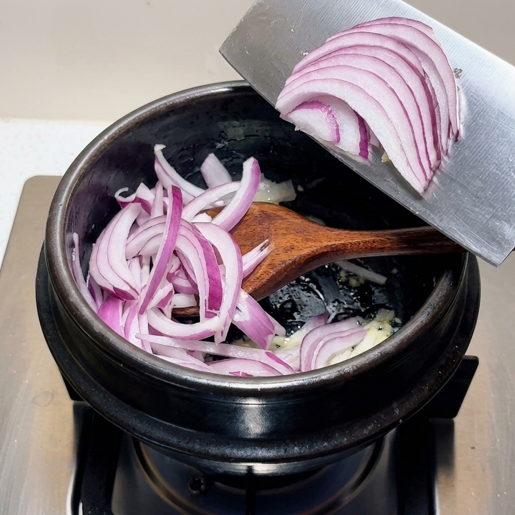 Una manciata di cipolle rosse viola affettate che vengono trasferite dalla lama di un grande coltello in una pentola nera con gli ingredienti aromatici.