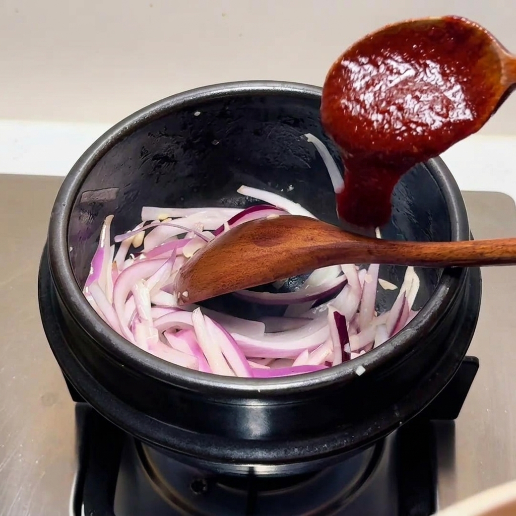 Un cucchiaio di legno che aggiunge una salsa densa e rossa di peperoncino alle cipolle rosse affettate in una pentola di pietra nera.