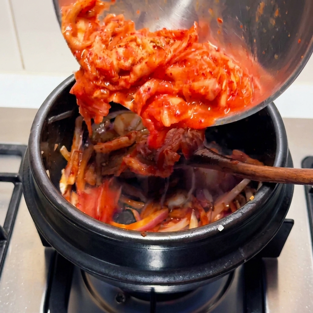 Kimchi aspro spezzettato di un rosso acceso che viene versato da una ciotola di acciaio inossidabile in una pentola di pietra nera sul fuoco.