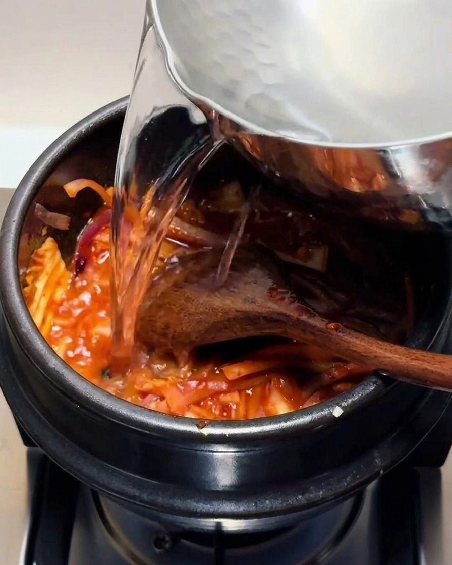 Acqua calda trasparente versata da un pentolino di metallo in una pentola di pietra nera contenente la miscela rossa di kimchi.