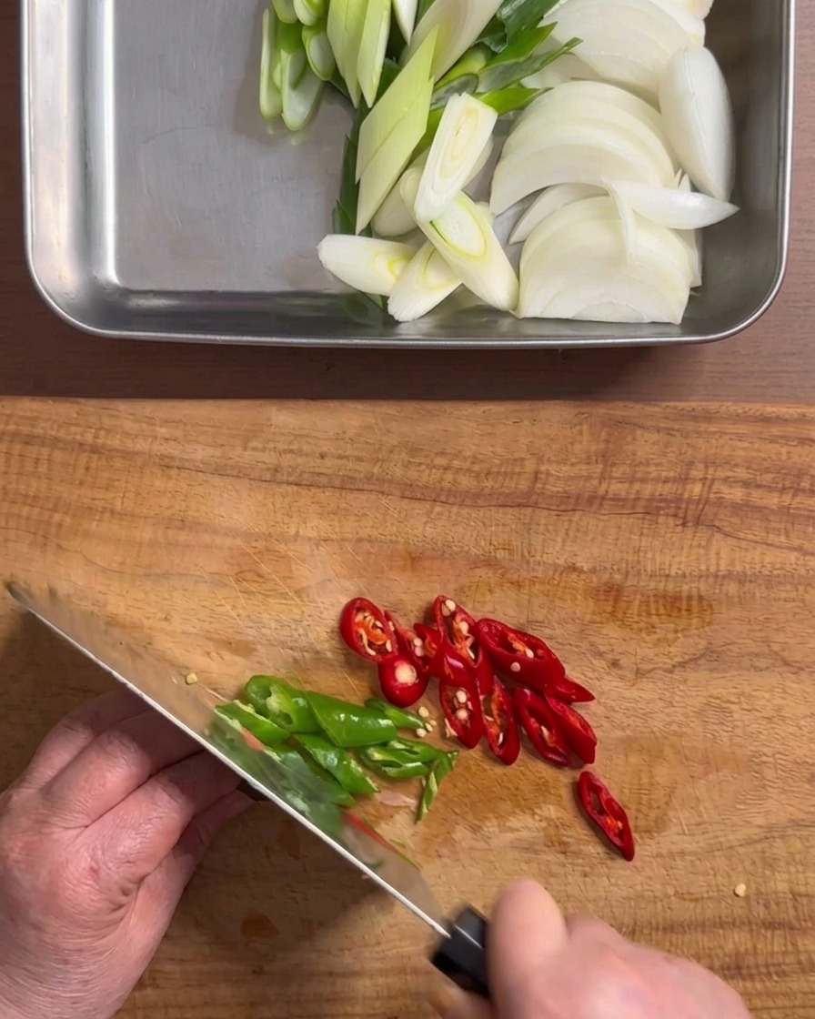 Mani che usano un coltello per tagliare peperoncini verdi e rossi a rondelle su un tagliere di legno.