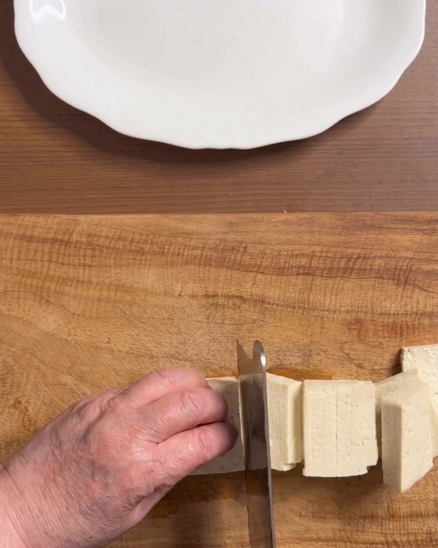 Mani che tagliano un blocco di tofu bianco sodo in pezzi rettangolari uniformi su un tagliere di legno.