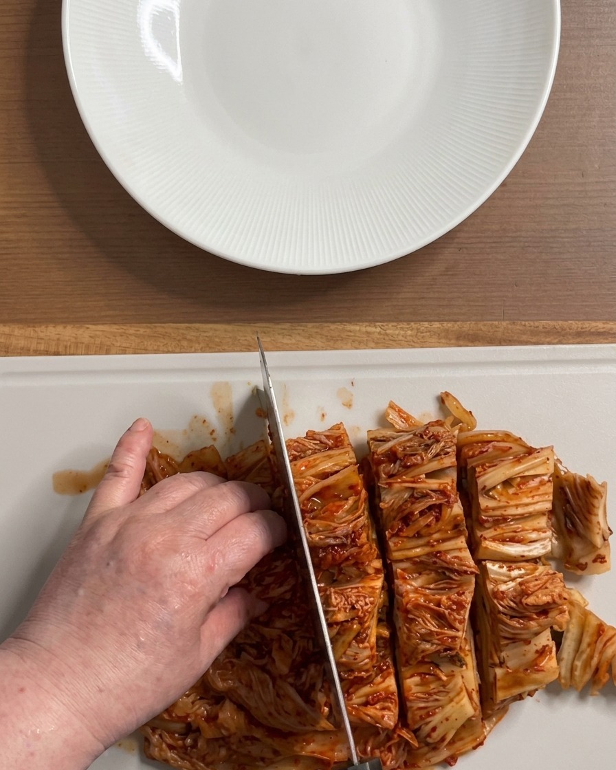 Mani che tagliano un grande blocco di kimchi rosso acceso a pezzi della grandezza di un boccone su un tagliere bianco.