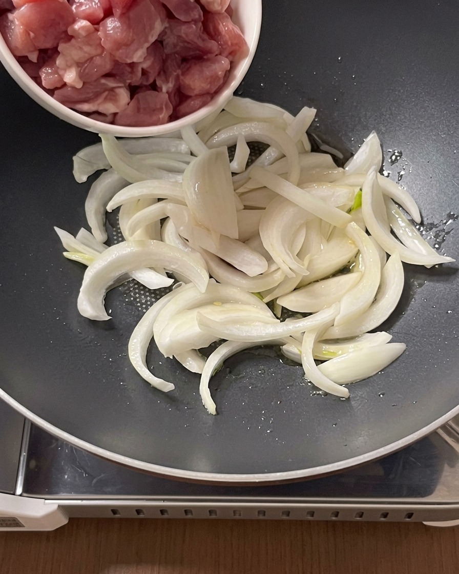 Pezzi di carne di maiale cruda aggiunti da una ciotolina in una padella con cipolle affettate e olio.