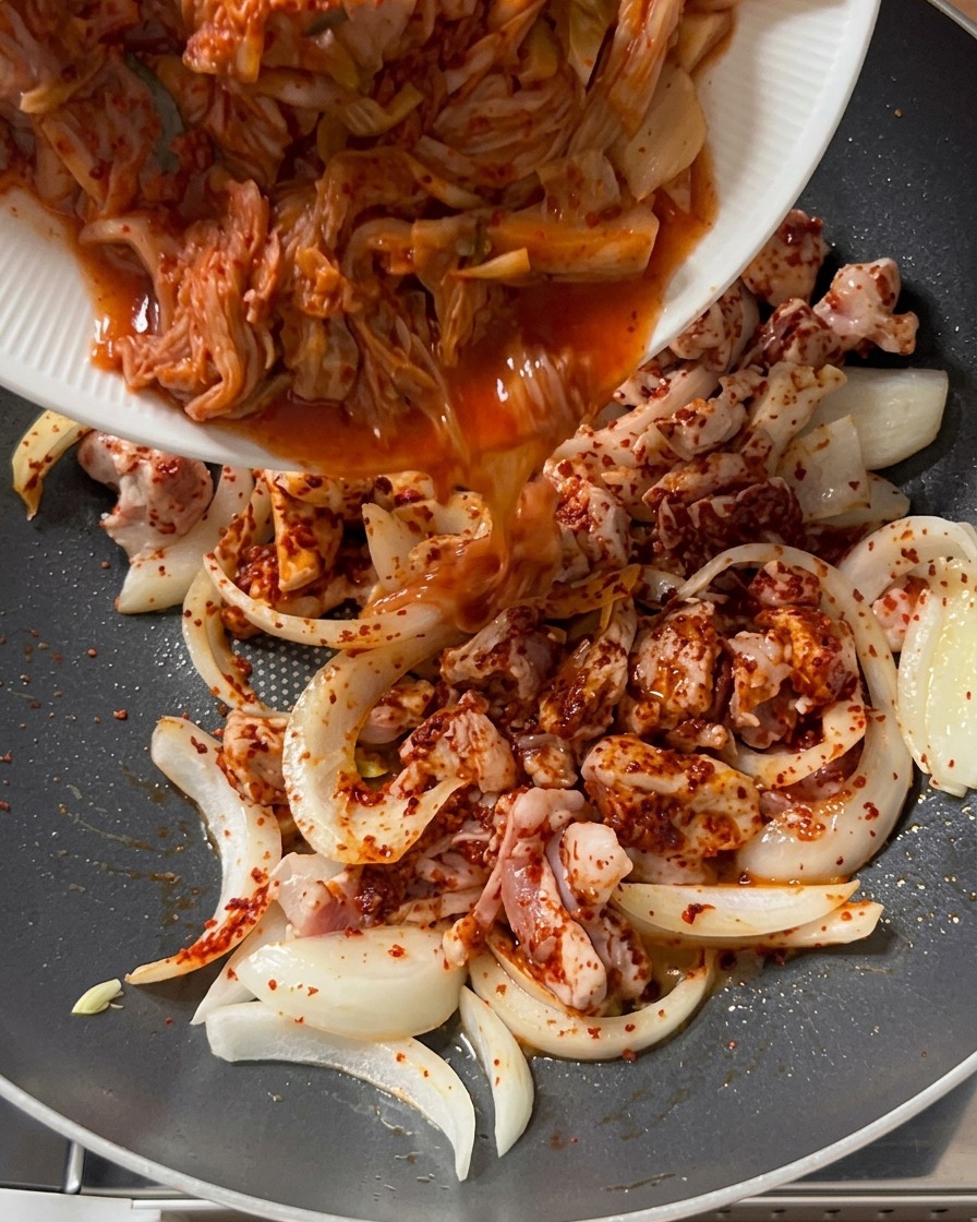 Kimchi spezzettato versato da un piatto bianco in una padella contenente maiale condito e cipolle.