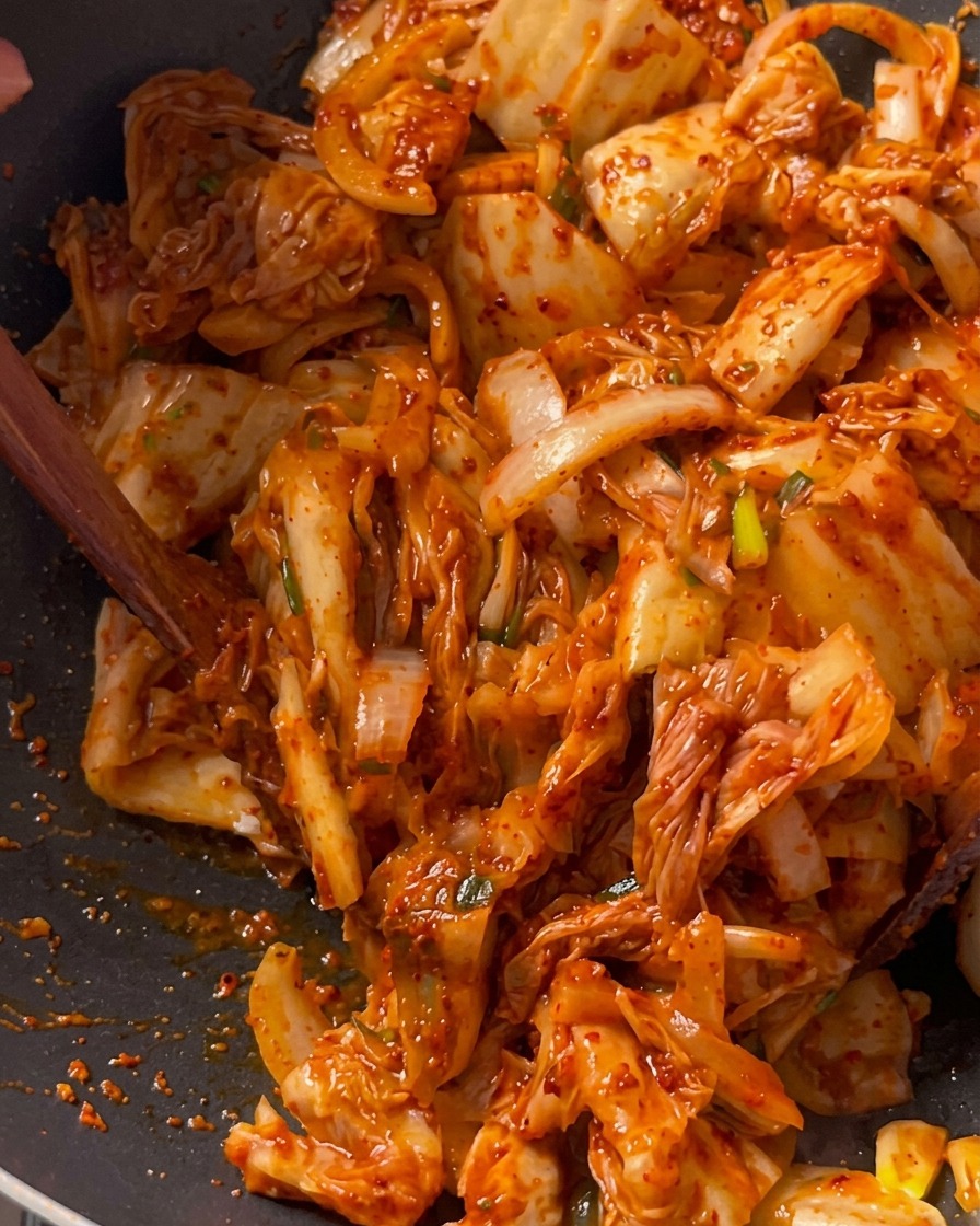 Pezzi di kimchi saltati insieme a carne di maiale e cipolle in un profondo wok nero usando una spatola di legno.