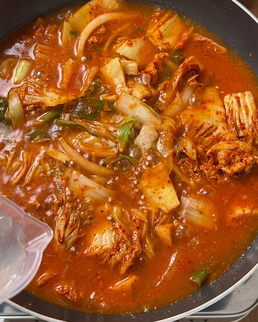 Acqua limpida versata da una caraffa di plastica trasparente in una grande pentola piena di un composto arancione-rossastro di kimchi e carne.