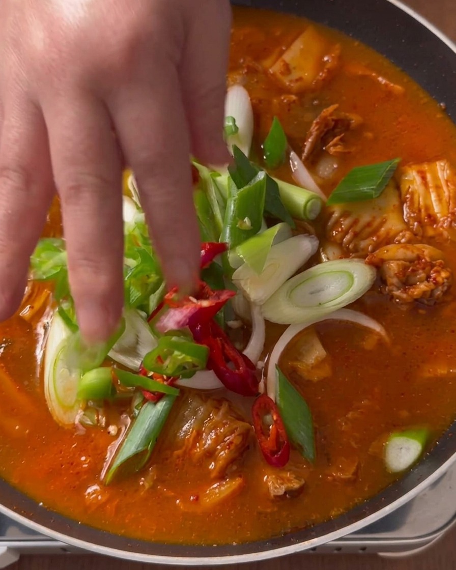 Una mano che adagia cipollotti freschi, peperoncini verdi e fette di peperoncino rosso sopra una zuppa di kimchi rossa che bolle in una pentola.