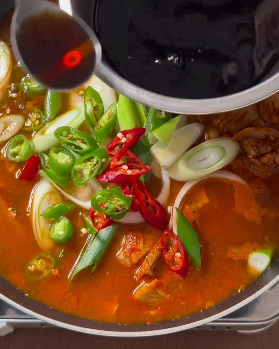 Un cucchiaio di condimento liquido scuro versato da una ciotola di acciaio inossidabile sopra una zuppa di kimchi guarnita con peperoncini freschi e cipollotti.