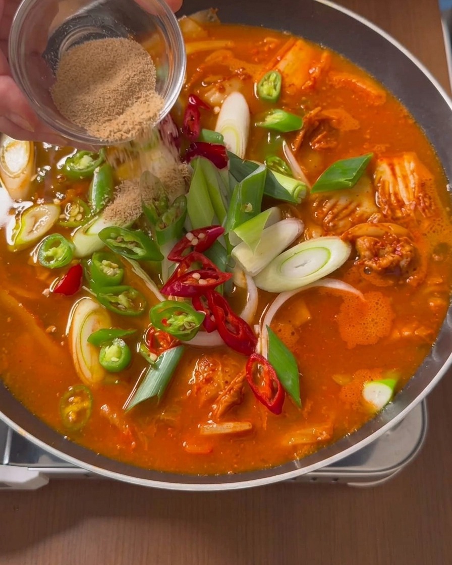 Insaporitore in polvere marrone spolverato da un piattino di vetro sulla superficie di una zuppa rossa in ebollizione con peperoncini e cipollotti.