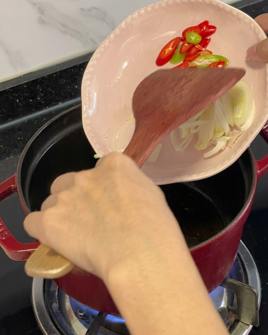 Peperoncini rossi e verdi affettati e aromi versati da un piattino rosa in una pentola rossa oliata usando una spatola di legno.