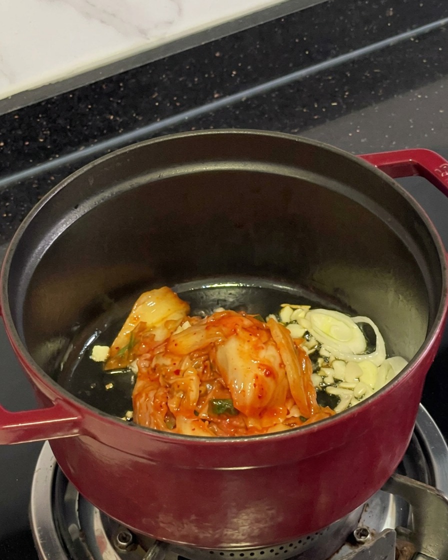 Kimchi rosso sminuzzato, aglio affettato e cipollotti che cuociono nellolio allinterno di una pentola.