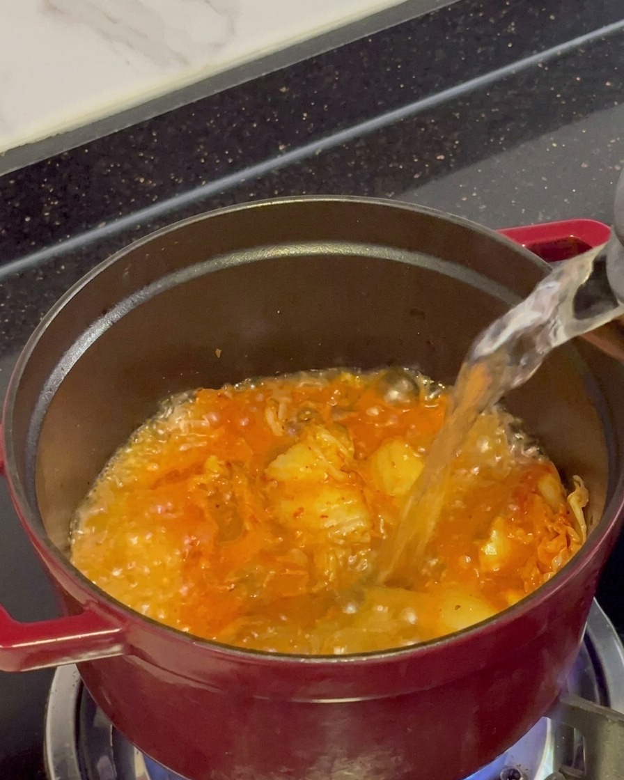 Acqua bollente limpida versata da un bollitore in una pentola rossa piena di kimchi che sfrigola.