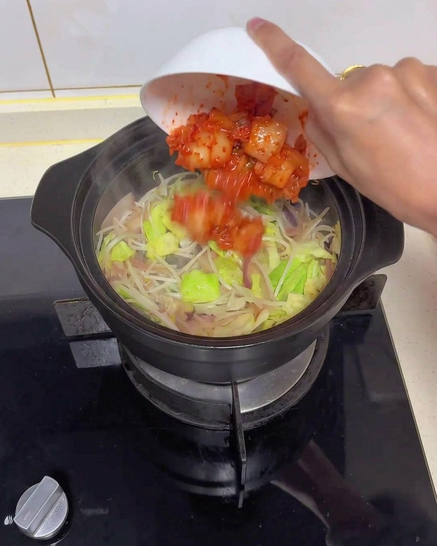 Una mano che inclina una ciotola bianca per aggiungere kimchi rosso a pezzi in una pentola di terracotta nera con cipolle, cavolo e germogli di soia.