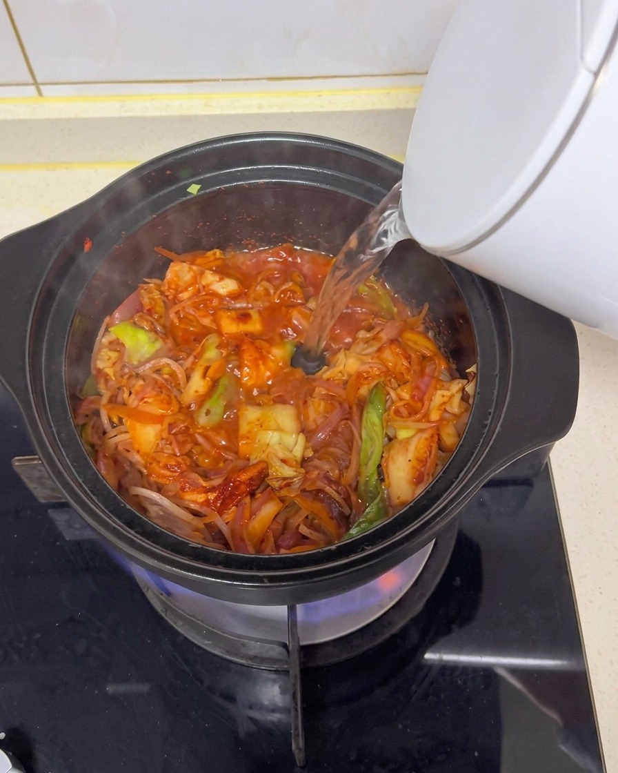 Acqua calda versata da un bollitore elettrico bianco in una pentola di terracotta nera su un fornello a gas, creando un brodo rosso con gli ingredienti saltati.