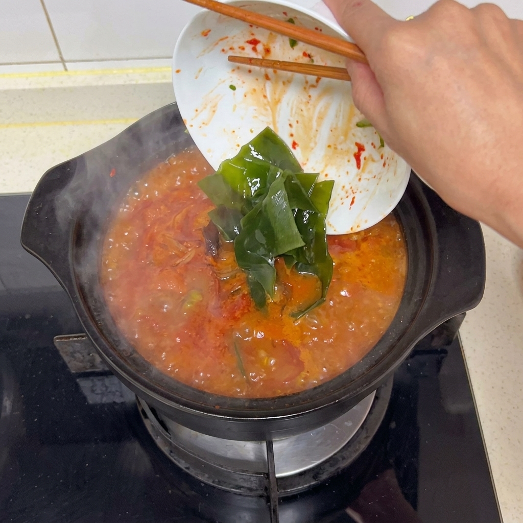 Bacchette che fanno scivolare ampie strisce di alga kelp verde scuro da una ciotola bianca in una zuppa rossa bollente.