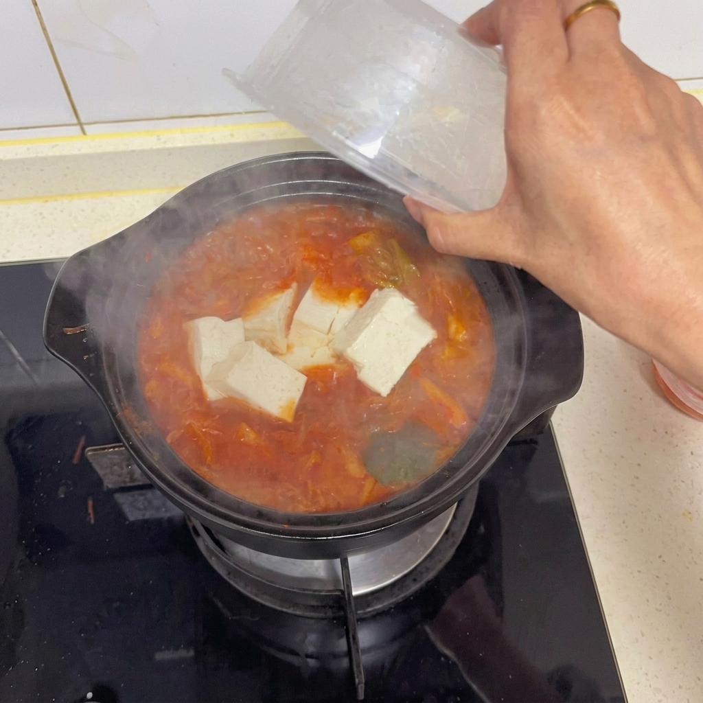 Una mano che aggiunge delicatamente grandi cubi di tofu bianco da un contenitore di plastica trasparente in una pentola di zuppa di kimchi rossa bollente.