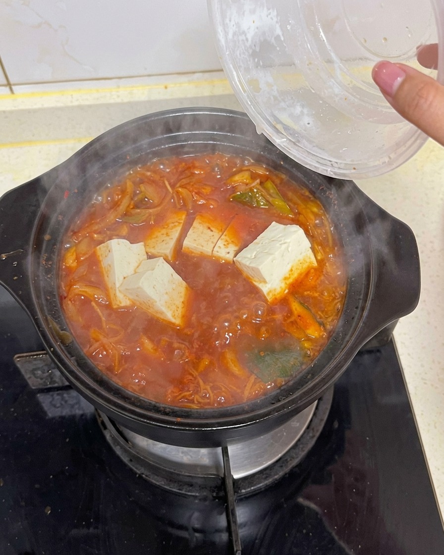Blocchi di tofu bianco rettangolare immersi da un contenitore trasparente in una pentola bollente di zuppa di kimchi rossa con verdure e alga.