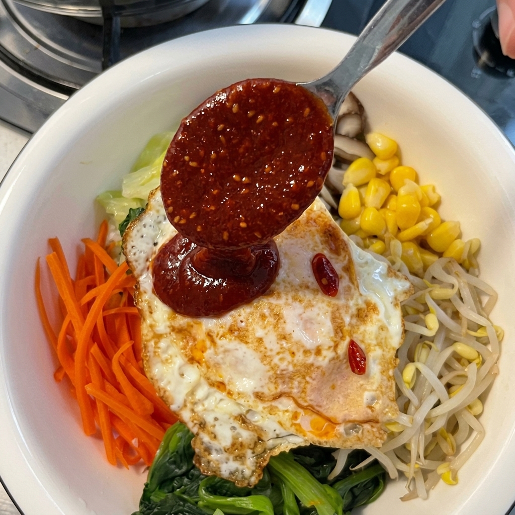 Salsa rossa al peperoncino versata da un cucchiaio di metallo sopra un uovo fritto adagiato su verdure per bibimbap.