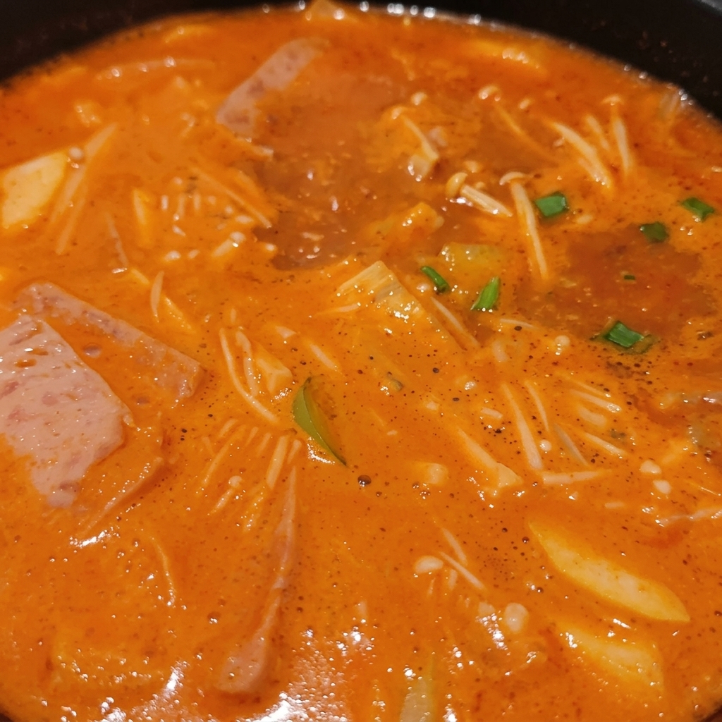 Uno stufato coreano bollente ricco di funghi enoki, fette di carne, cipollotti verdi e tofu immersi in un denso brodo arancione.