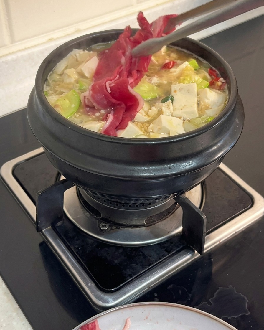 Pinze da cucina che adagiano sottili fette di petto di manzo crudo in una pentola di zuppa coreana con tofu e zucchine.