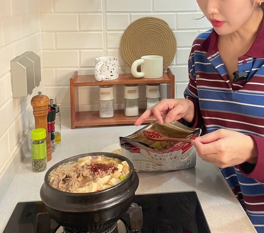 Un cucchiaio di peperoncino rosso a grana grossa aggiunto a una pentola di terracotta con zuppa coreana di soia, tofu e carne.