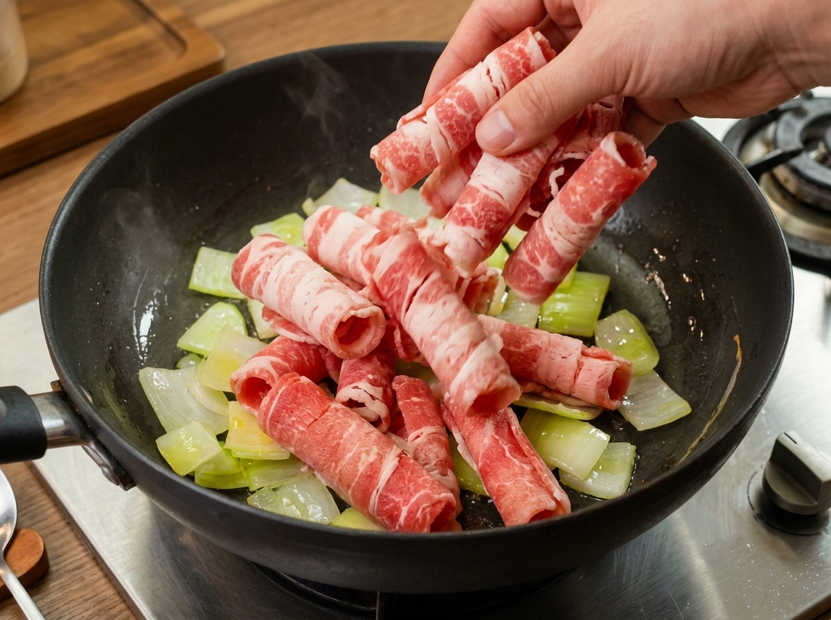 Sottili fette di manzo crudo arrotolate aggiunte in una pentola con cipolle saltate traslucide.