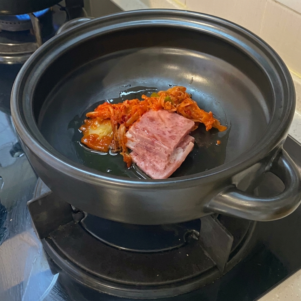 Fette di pancetta di maiale cruda e vibrante kimchi tritato nellolio sul fondo di una pentola di terracotta scura.