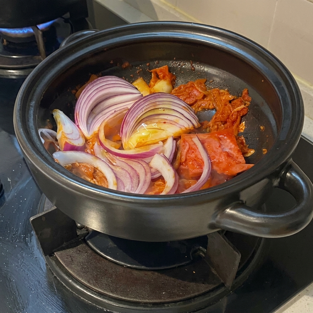 Spesse fette di cipolla rossa adagiate sopra il maiale e il kimchi saltati allinterno di una pentola di terracotta.