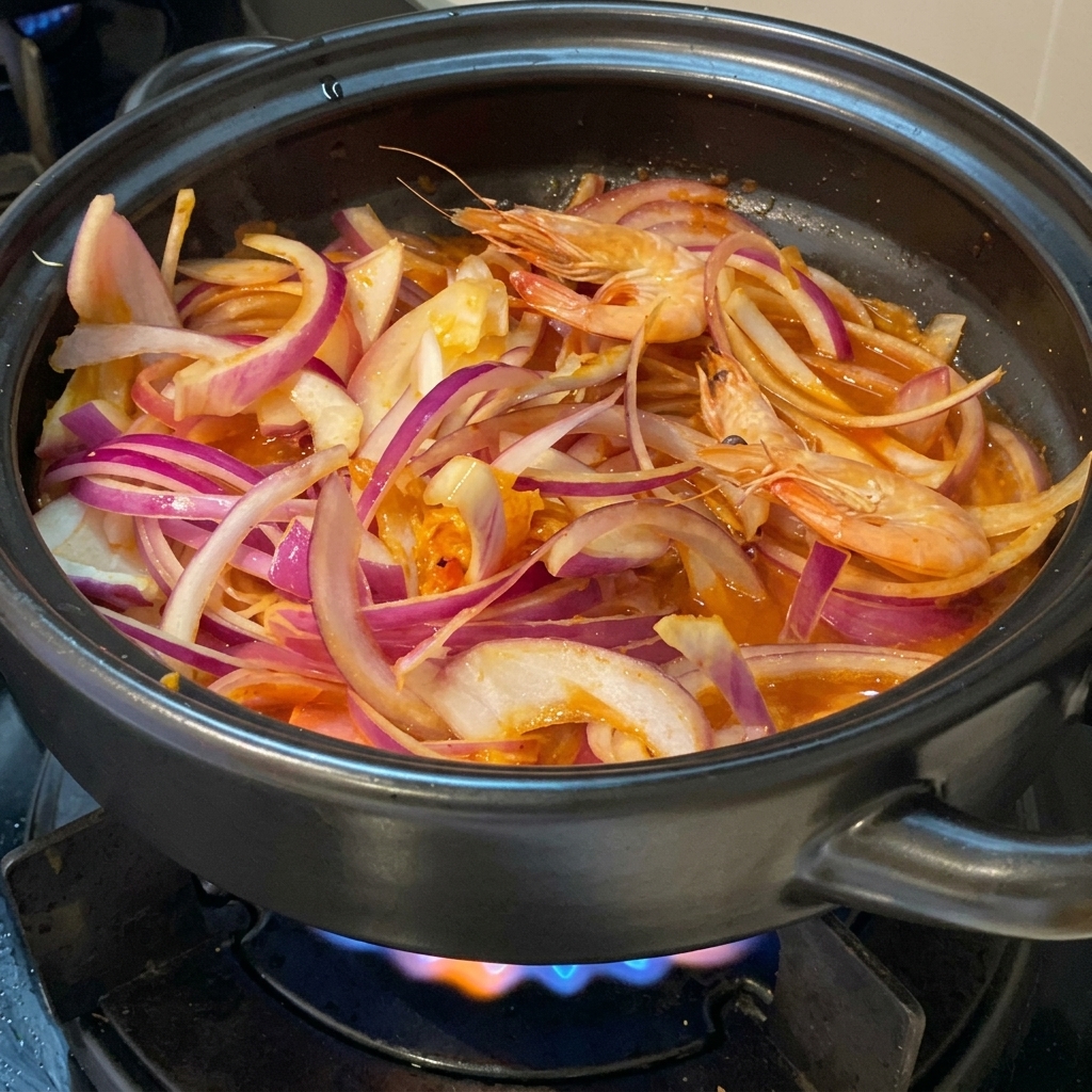Gamberi rosa freschi interi adagiati tra fette di cipolla rossa e kimchi in una pentola di terracotta nera su un fornello acceso.