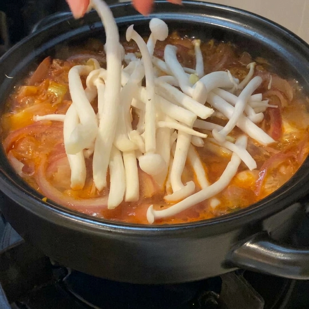 Funghi di mare bianchi e freschi disposti con cura a mano in una zuppa di kimchi rossa e spumeggiante dentro una pentola di terracotta nera.