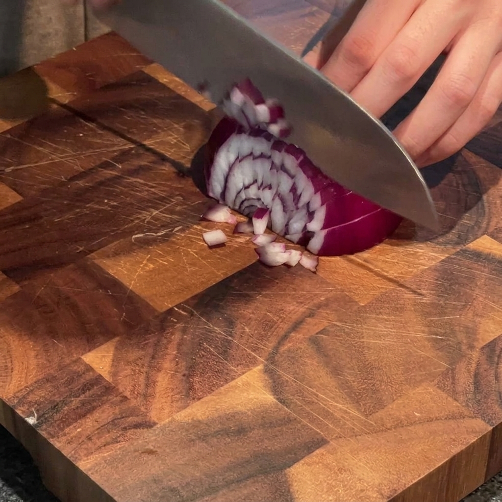 Una mano che usa un coltello da chef per tritare una cipolla rossa a piccoli pezzi su un tagliere di legno.