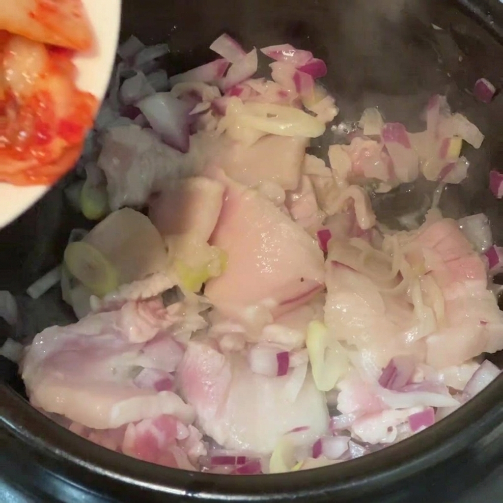 Kimchi a pezzi aggiunto da un piatto in una pentola con pancetta di maiale e cipolle rosse.