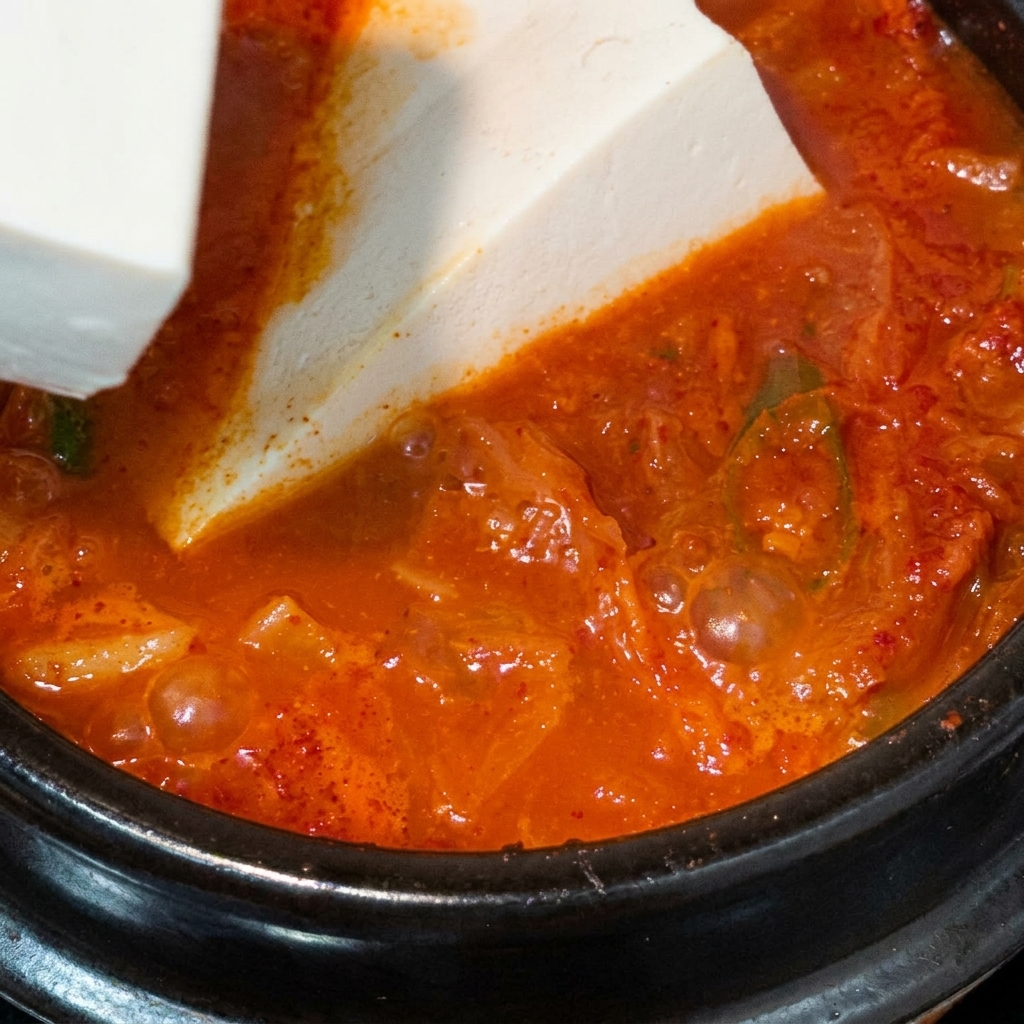Un grande blocco di tofu vellutato bianco posato con cura in una pentola bollente di zuppa di kimchi rossa.