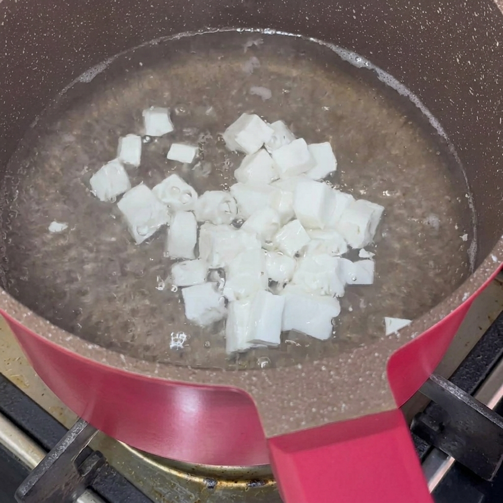 Piccoli cubetti di tofu bianco morbido che sbollentano in acqua in una pentola antiaderente rosa.