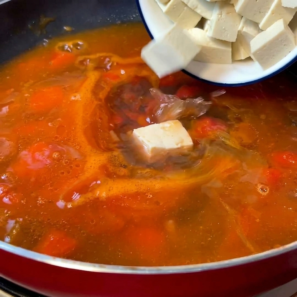 Cubetti bianchi di tofu delicatamente aggiunti in una pentola con la base di zuppa al pomodoro bollente.
