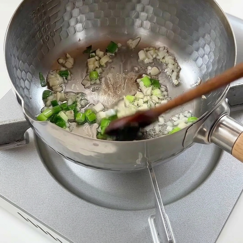Cipollotti tritati e aglio tritato rosolati in olio caldo allinterno di una pentola di metallo.