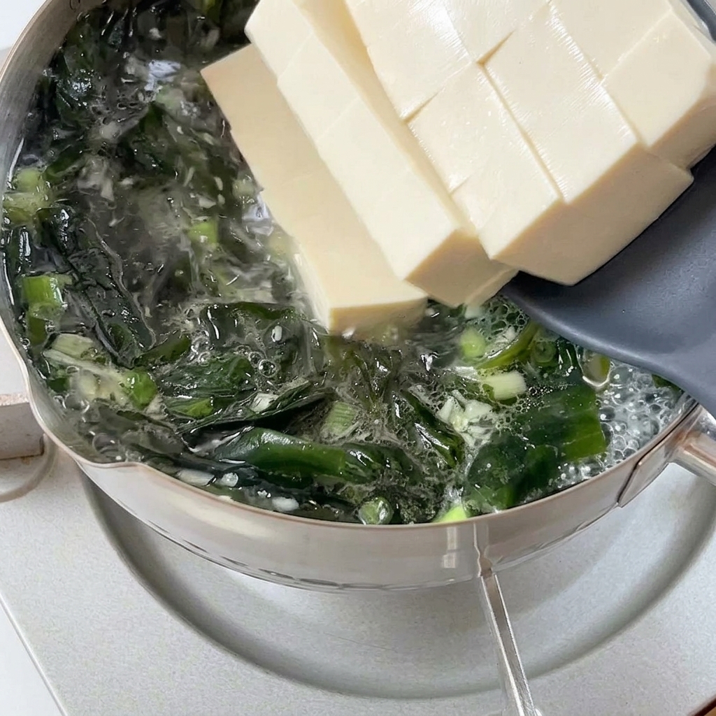 Spesse fette di tofu bianco e morbido fatte scivolare da una spatola in una pentola fumante di brodo con wakame.