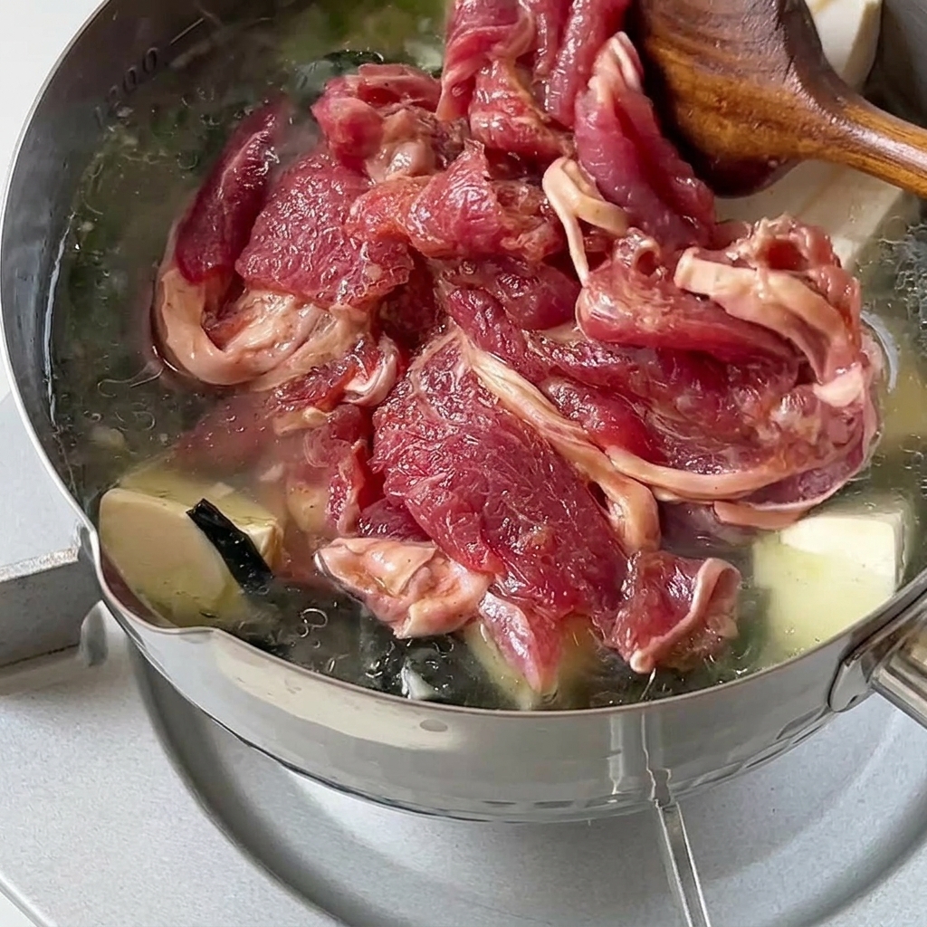 Fette di manzo crudo marinato aggiunte delicatamente e mescolate in una pentola calda di zuppa con wakame e tofu.