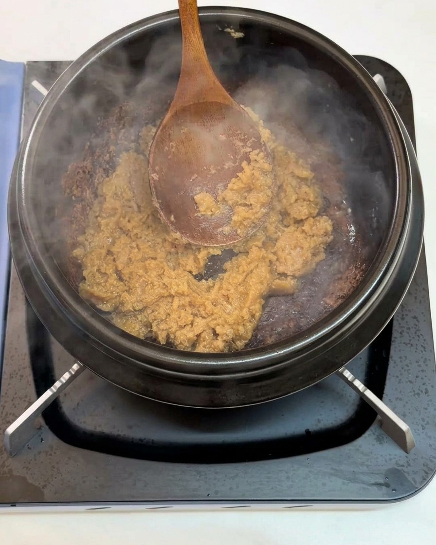 Un cucchiaio di legno che mescola aglio tritato e pasta di soia in un tegame nero di terracotta sul fuoco.